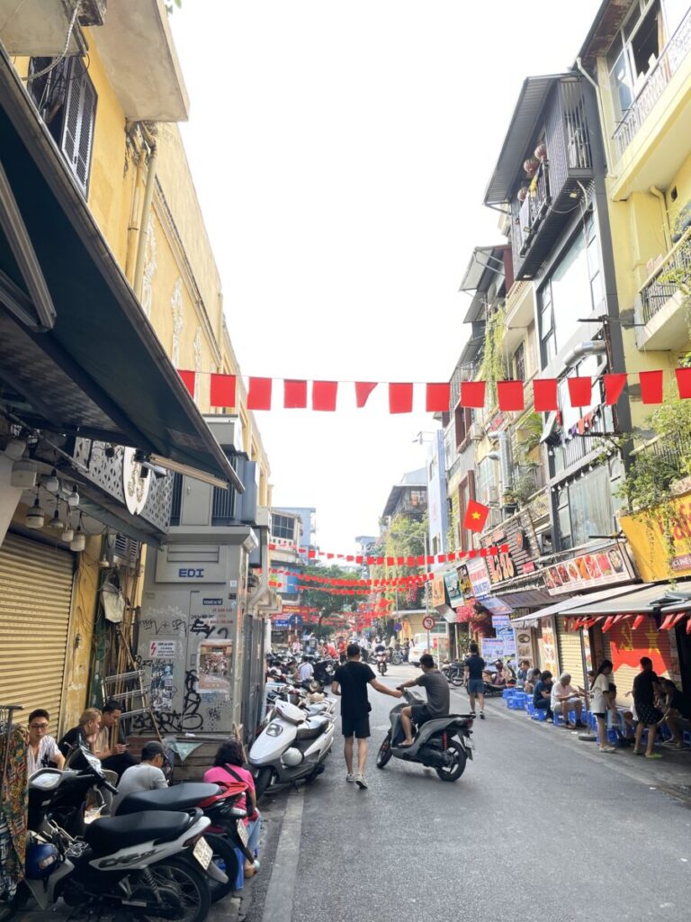 ハノイ旧市街の街並み。赤いベトナム国旗が連なる通りとローカル食堂、バイクが行き交う昼の風景