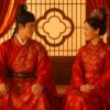 中国の伝統婚礼衣装をまとった新郎新婦が、花窓を背景に向かい合って座っている。窓越しの格子と柔らかな光が、厳かで華やかな雰囲気を引き立てている。