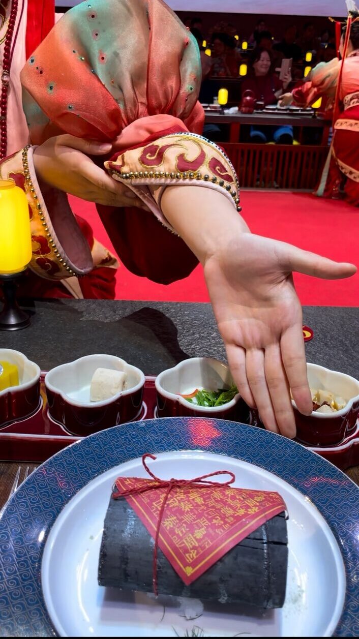 炭のように黒い竹筒風の料理を小さな金づちで割る演出。周朝風衣装の給仕スタッフの手元と赤い小皿が並ぶ宮廷ディナーの一場面。