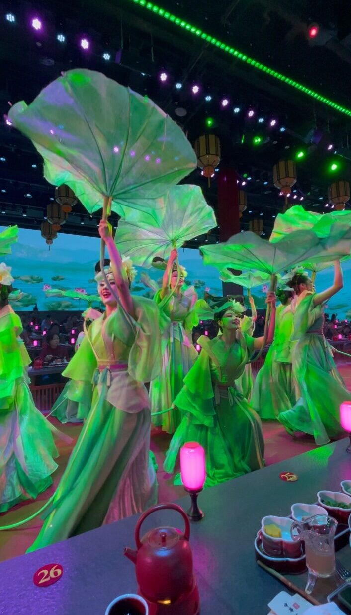 北京御仙都皇家庭菜の宮廷ディナーショーで、緑の衣装をまとった舞姫たちが大きな蓮の葉を掲げて舞う幻想的なシーン。