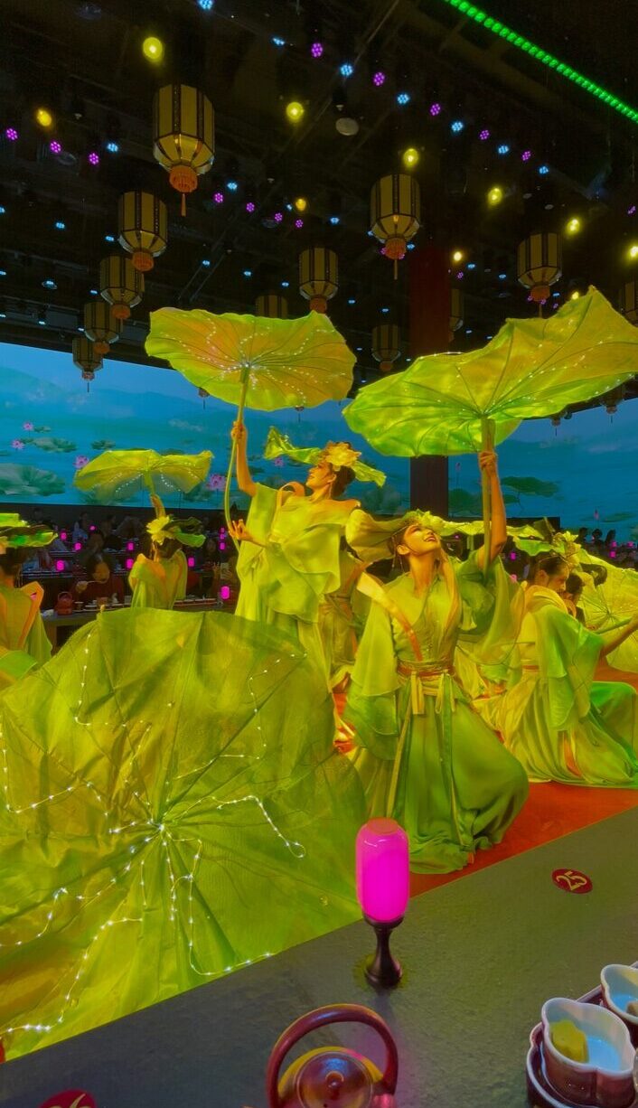 北京の宮廷ディナーショーで、緑の衣装をまとった舞姫たちが光をまとった大きな蓮の葉を掲げて舞う幻想的な演出シーン。