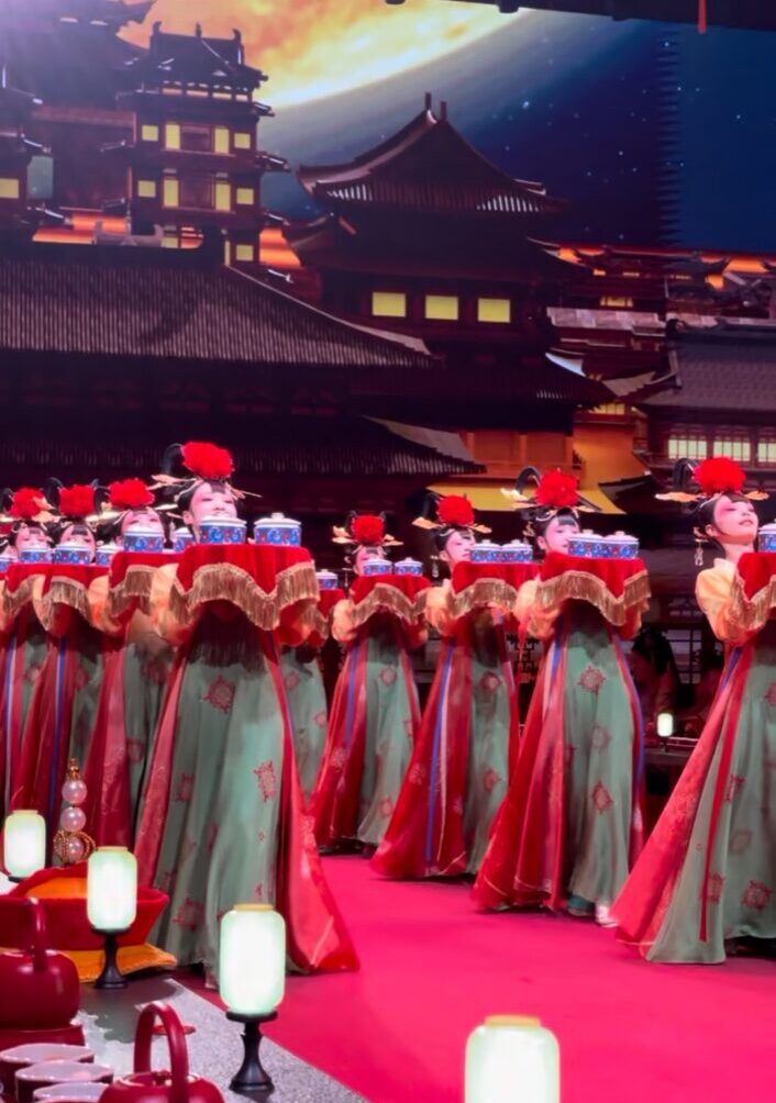 宮廷衣装の女性たちが料理を高く掲げ、上体を大きく反らせながら進む献上風の演出。赤い舞台と宮殿背景の中で整然と歩む所作が印象的。