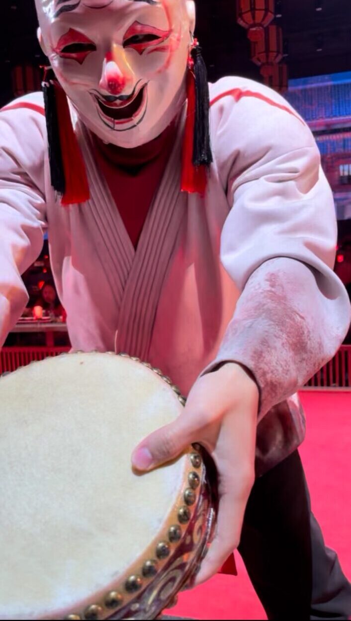 道化風の仮面をつけた演者が、小さな太鼓を観客に差し出す場面。赤い照明の舞台でコミカルな演出が行われている様子。