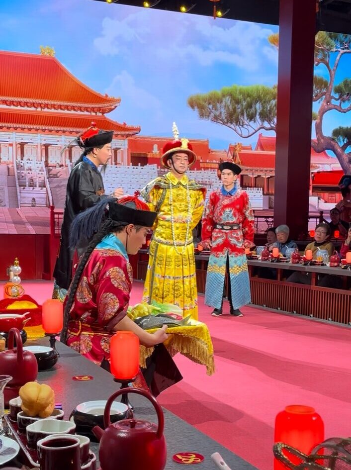 紫禁城を模した背景の舞台で、黄色の皇帝衣装をまとった人物を中心に、清朝風の衣装を着た演者たちが儀式の場面を演じている様子。客席のすぐ前で宮廷劇が展開されている。