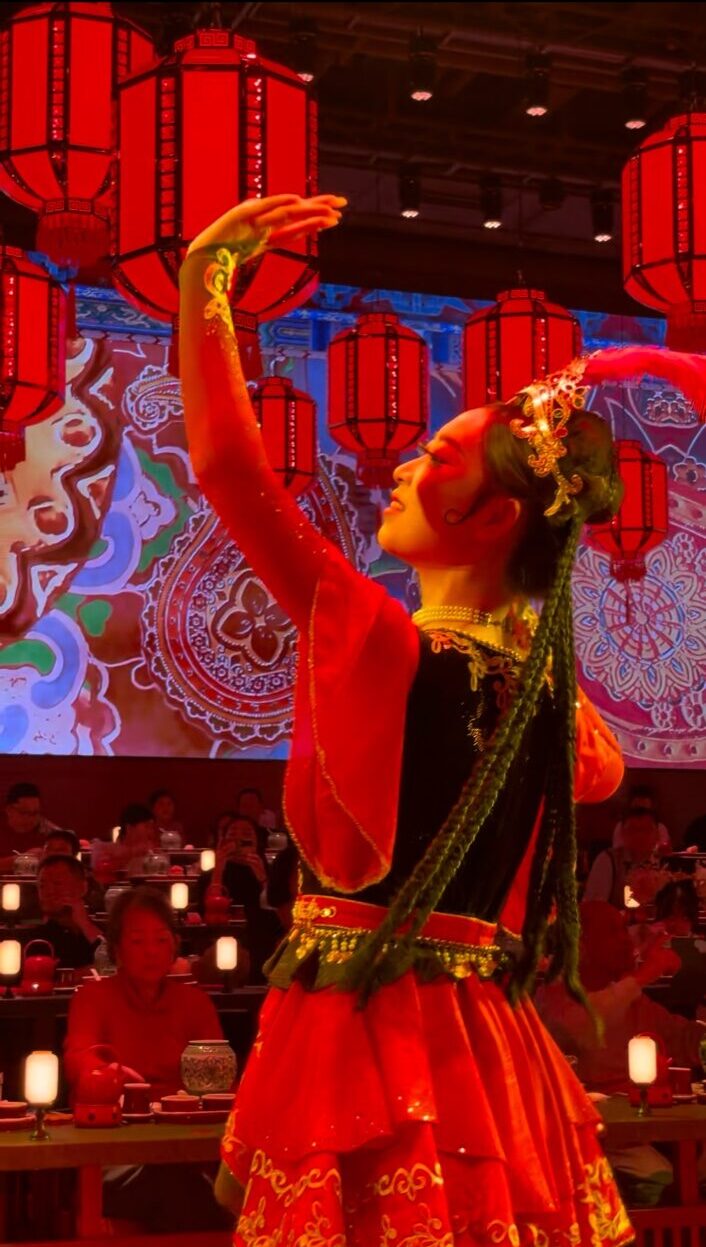 赤い衣装をまとった女性ダンサーが、赤い提灯が吊るされた舞台の前で優雅に腕を伸ばして舞っている様子。背景には中国風の文様と観客席が広がっている。