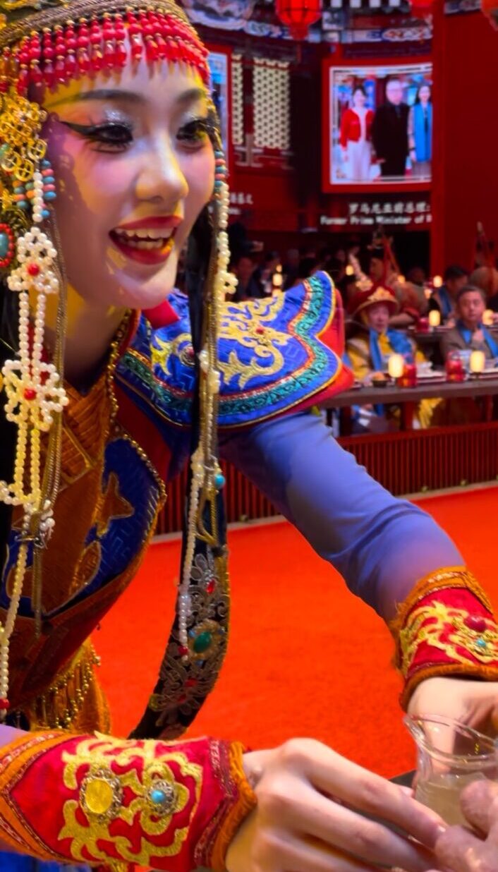 華やかな民族衣装をまとった女性ダンサーが、笑顔で客席に飲み物を差し出すパフォーマンス。