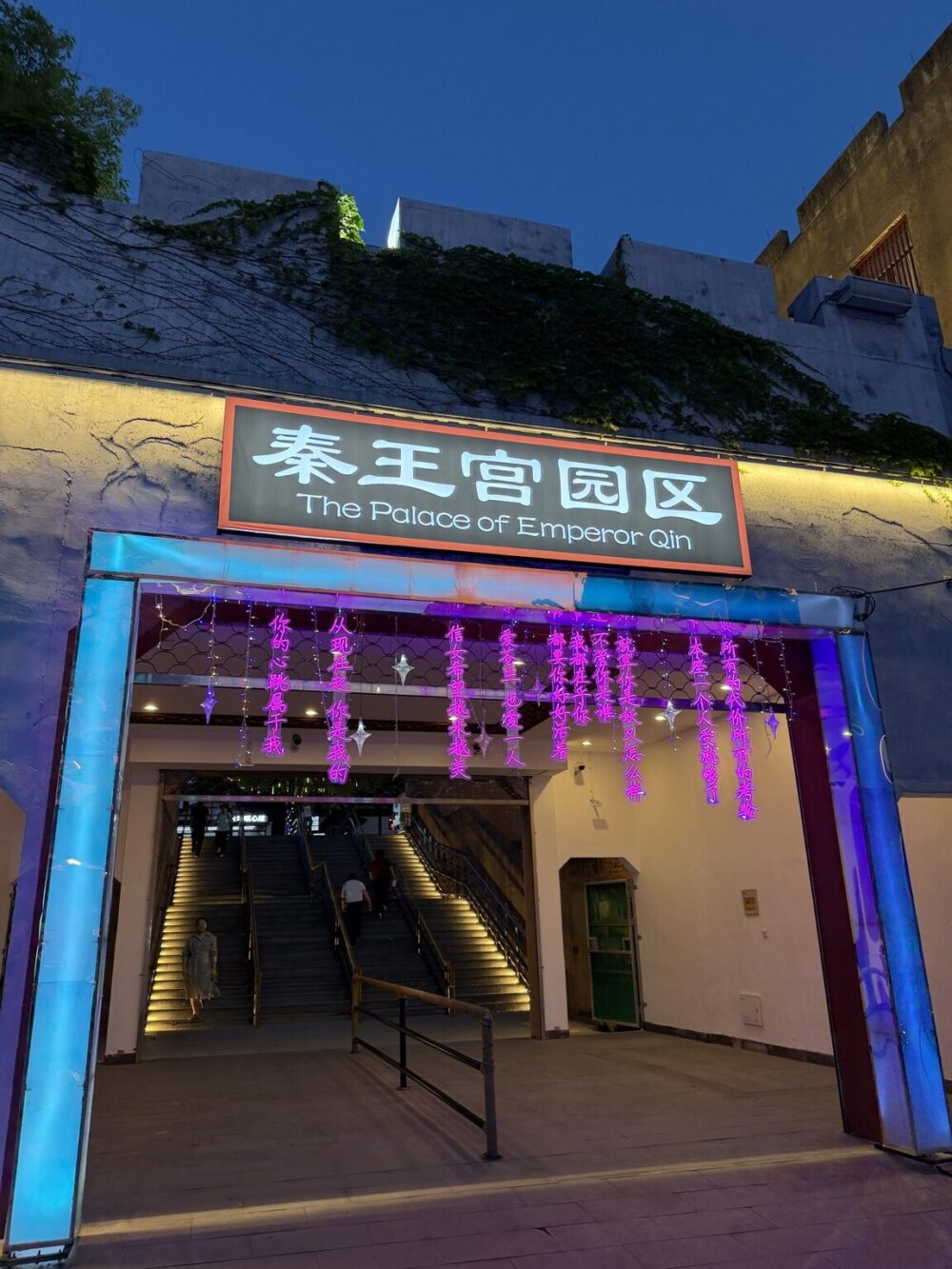 清明上河図景区内に設けられた秦王宮園区への連絡通路入口。夜のライトアップに照らされた「秦王宮園区（The Palace of Emperor Qin）」の看板と、上階へ続く階段。