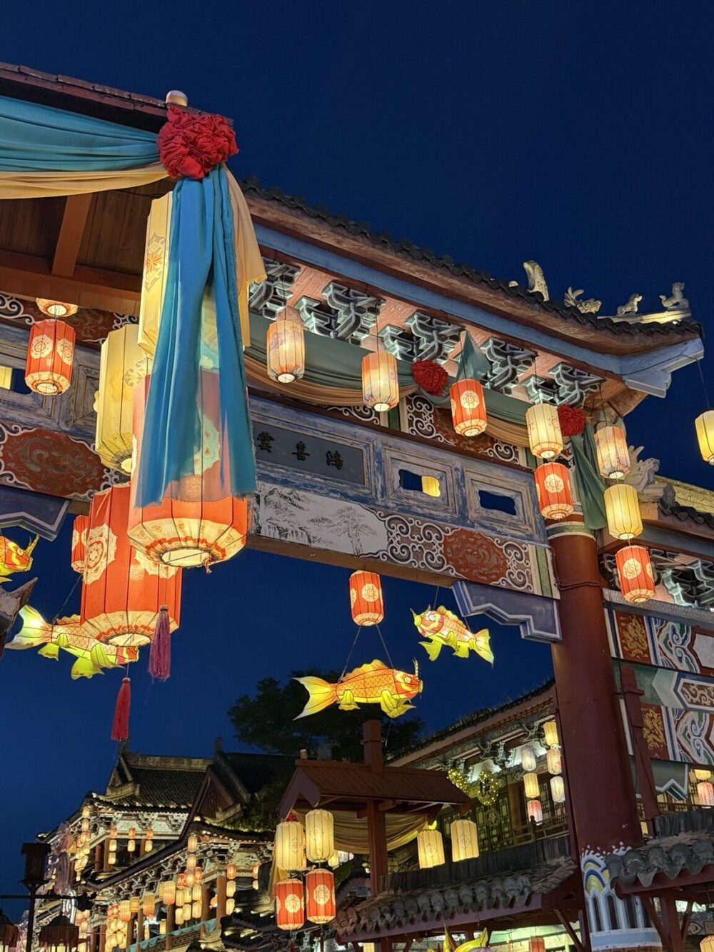 横店影視城・清明上河図の夜景。提灯が灯る門に色鮮やかな魚の花灯が飾られ、北宋の街並みを華やかに彩る様子 キャプション